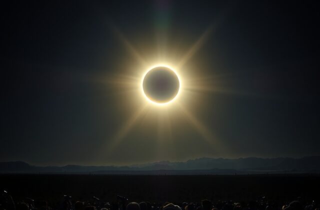 A wide-angle shot of a total solar eclipse over a scenic landscape with a silhouetted crowd wearing eclipse glasses, the sun’